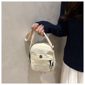 Detail of a beige pleated crossbody bag with a round metal logo and wide textile strap