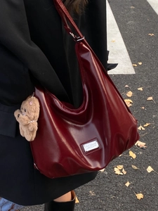 Large burgundy patent leather hobo bag with plush bear charm on autumn street
