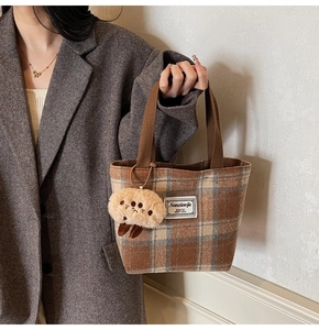 Detail of a brown plaid tote bag with a white brand label and a plush doll charm