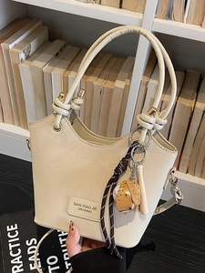 Chic creamy white bucket bag with knot handle and baseball charm on a bookshelf