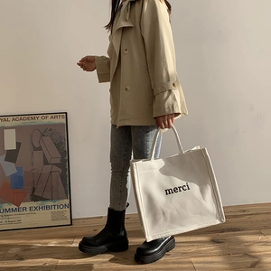 White canvas tote bag with black merci print styled with a classic beige trench coat