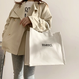 Detailed shot of white canvas tote bag with black merci print on a beige trench coat