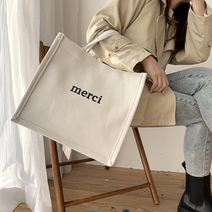 Detail of a beige canvas tote bag featuring black merci print in a lifestyle setting