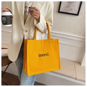 Close-up of a yellow canvas tote bag with black merci lettering held by a person