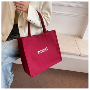 Detail of burgundy canvas tote bag with white merci text held by a model in white blazer