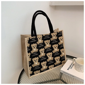 Detail of jute tote bag featuring cartoon bear print and black braided handles on glass table