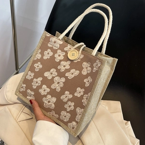 Square jute woven handbag with brown panel, white floral pattern, and wood button detail