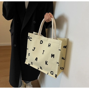 Detail of a beige canvas tote bag with black letter print held by a model in a black coat