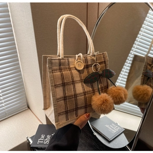 Detail of plaid tote bag featuring thick rope handles, wooden button, and cherry pom pom