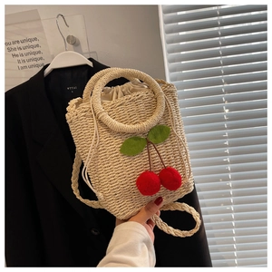 Detail of beige straw bucket bag featuring plush red cherries and a woven shoulder strap