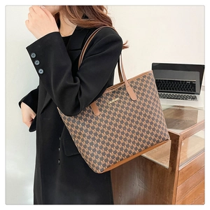 Detail of a large brown monogram tote bag featuring a gold metal nameplate and PU leather texture