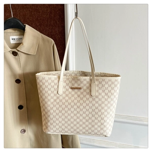 Detail of cream tote bag with M-pattern monogram and gold metal plaque on a trench coat