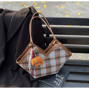 Detail view of plaid tote bag with brown leather straps and teddy bear charm on bench