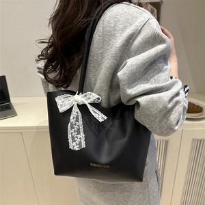 Minimalist black tote bag with white lace bow and gold logo shown on model's shoulder