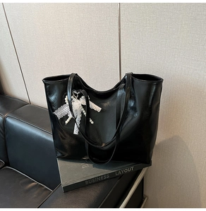 Detail of glossy black tote bag with white lace bow and pearl pendant on leather sofa