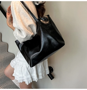 Model wearing a large black high-gloss tote bag over the shoulder in a minimalist setting