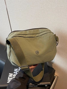 Olive green nylon crossbody bag with wide adjustable strap and metal logo on beige background