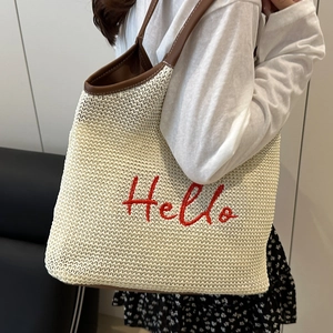 Woven straw tote bag with brown leather straps and red Hello embroidery on white texture