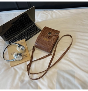 Brown woven leather crossbody phone bag on a white bed with modern lifestyle props