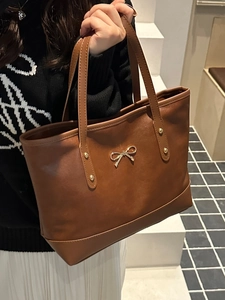 Brown leather tote bag featuring a gold bow ornament and rivets in a modern interior setting