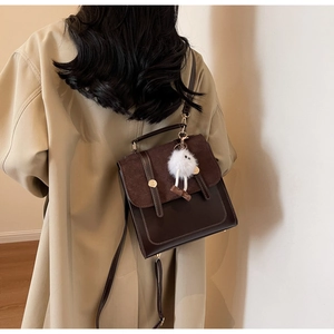Brown leather crossbody bag with plush charm shown on a model in a beige trench coat