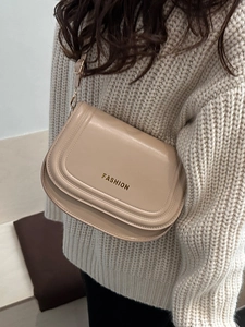 Chic beige saddle crossbody bag with gold Fashion logo on a soft white knit sweater