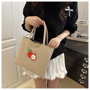 Detail of embroidered red apple pattern on beige nylon tote bag held by a model