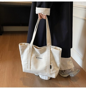 Detail of a beige canvas tote bag with BE YOURSELF print and outer pocket design