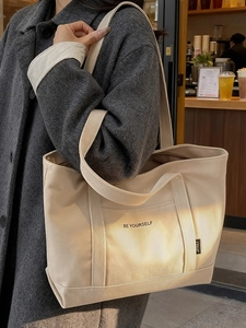 Beige canvas tote bag with black Be Yourself lettering shown on a model shoulder