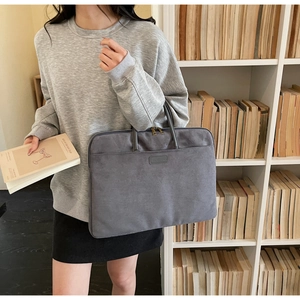Grey suede laptop bag with dual handles and leather logo patch shown in office setting
