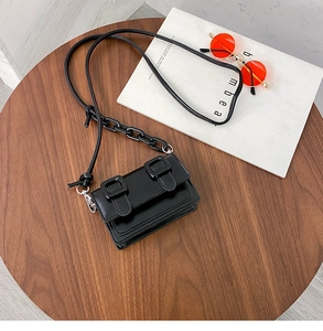 Detail of a black mini crossbody bag with square buckles and chain strap on a wooden table