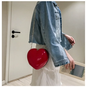Red heart-shaped patent leather bag with gold chain strap worn by a model in casual setting