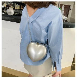 Silver heart-shaped mini chain bag shown held by a model in a cafe setting