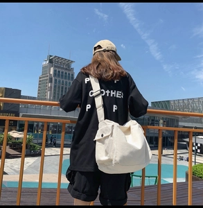 Outdoor street style view of a large cream canvas messenger bag with a black lettered T-shirt