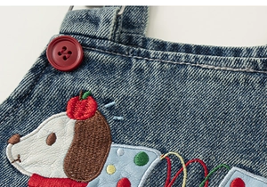 Detail of blue denim overalls featuring a red button and cute puppy embroidery for kids