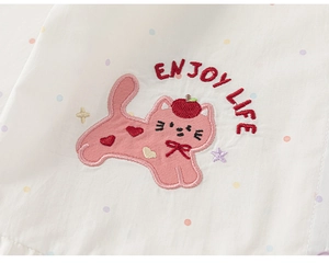 Dettaglio del ricamo di un gatto rosa con testo Enjoy Life su camicia in cotone bianco