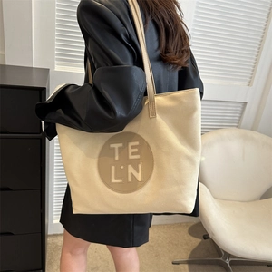 Detail shot of a beige canvas tote bag featuring a leather logo patch and shoulder straps