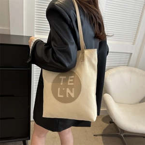 Detail of beige canvas tote bag with TE LN leather patch worn with a black leather jacket