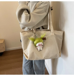 Close-up of beige corduroy tote bag featuring a green plush doll pendant and long shoulder straps