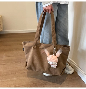 Detail of a brown corduroy tote bag featuring a plush doll charm and leather logo patch