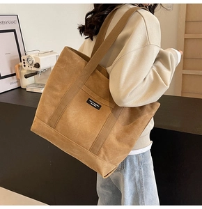 Side view of a large caramel canvas tote bag with black label worn by a model in a cafe