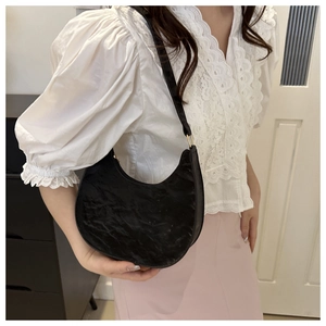 Detail of a black pleated PU leather saddle bag held by a model in a casual indoor setting