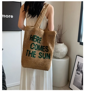 Detail of woven straw tote bag with green embroidered Here Comes The Sun text