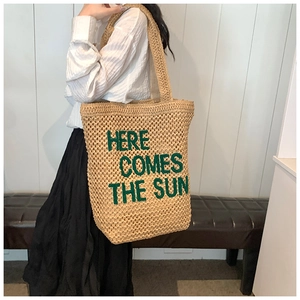 Detail of straw tote bag featuring green 'HERE COMES THE SUN' embroidery on woven texture