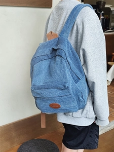 Close-up of denim backpack with brown leather logo patch worn by a model in a modern room