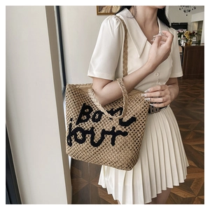 Detail of a beige woven straw tote bag with black Bonjour embroidery and braided handles
