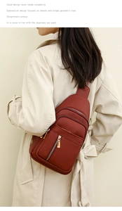 Brick red crossbody chest bag with pebble leather and gold zippers worn over a beige coat