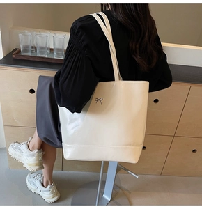 Detail of a creamy white glossy PU tote bag featuring a delicate metal bow ornament