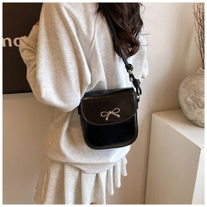 Detail of a black glossy saddle bag featuring a gold bow accent and knotted shoulder strap