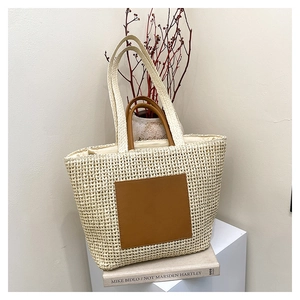 Large beige woven straw tote bag with brown leather patch on a minimalist pedestal